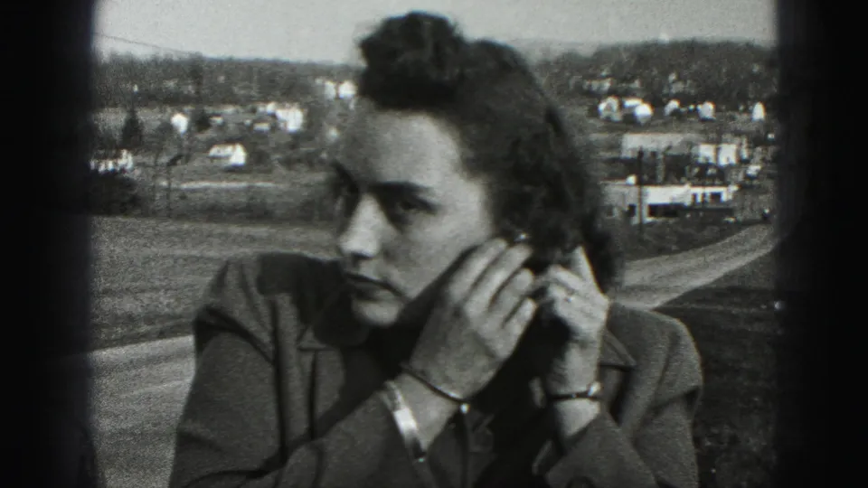 The woman continues fixing her earring in close view, framed against open ground and distant houses near Harrisburg, Pennsylvania.