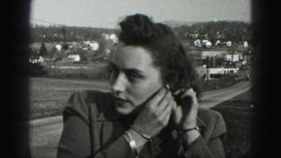 In 1975 Harrisburg-area 8mm footage, a woman in a jacket adjusts an earring beside a road with houses in the distance.