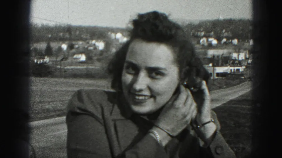 A woman in 1975 Harrisburg footage smiles toward the camera while adjusting her earring against a suburban roadside background.