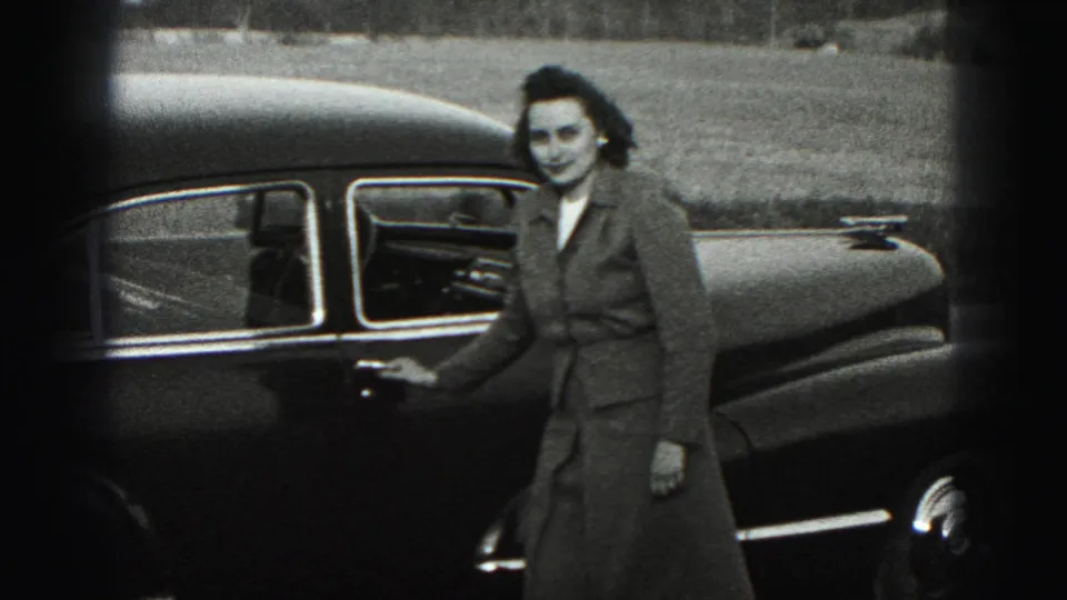 In Harrisburg, Pennsylvania, a woman in a mid-1970s coat stands by a dark sedan in grainy black-and-white 8mm footage.