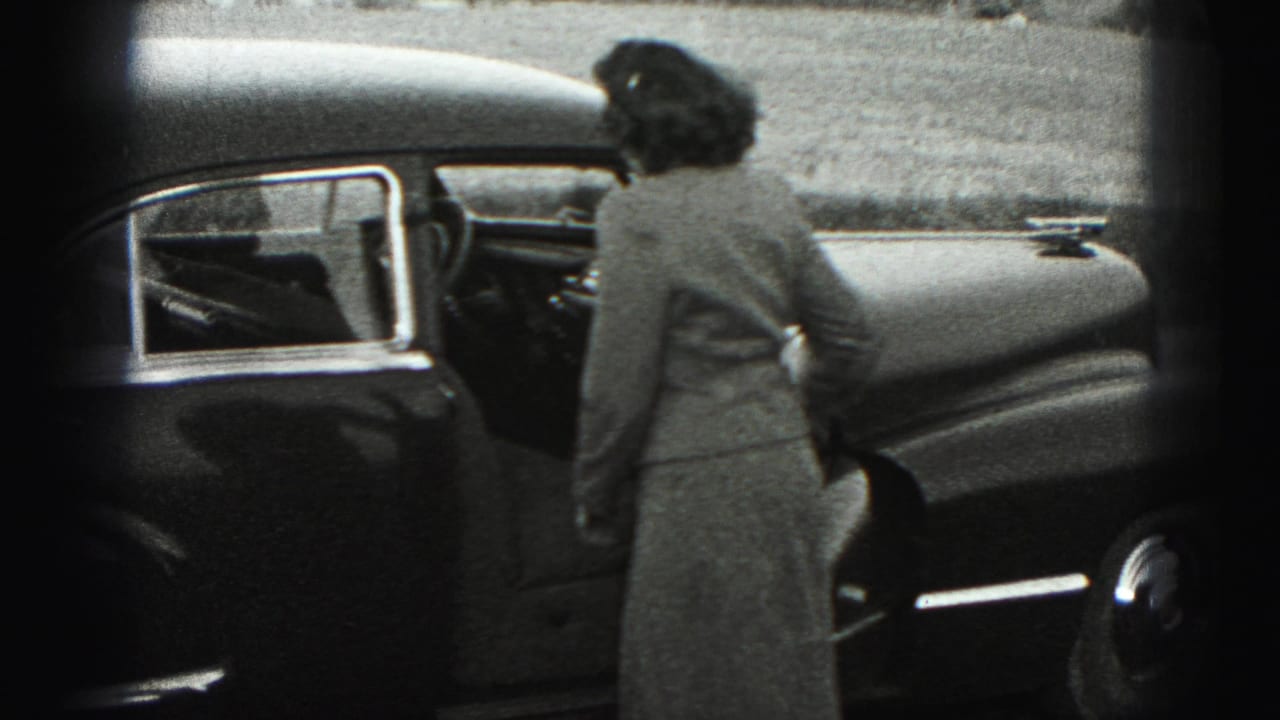 The woman turns away from the camera and opens the driver's door of the sedan in the Harrisburg home-movie scene.