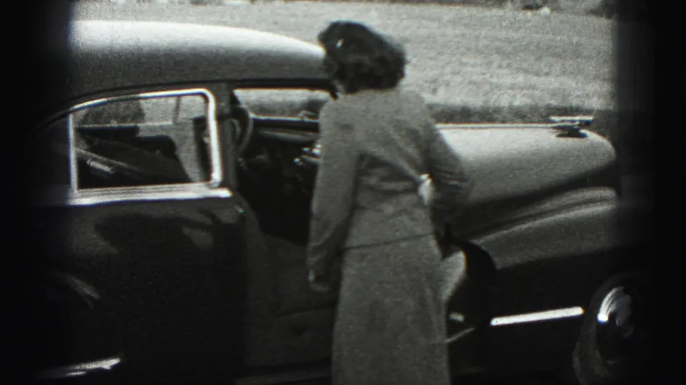 The woman turns away from the camera and opens the driver's door of the sedan in the Harrisburg home-movie scene.