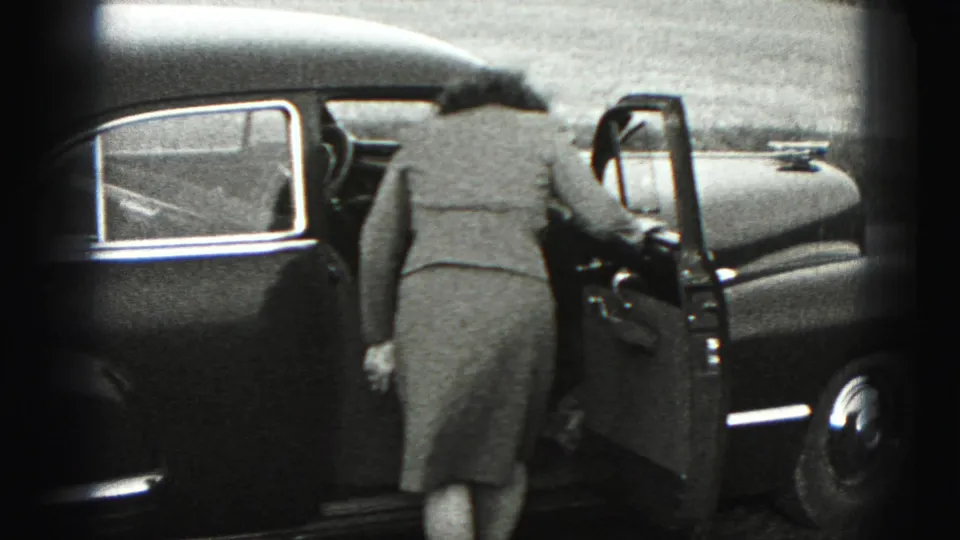 A woman in a coat stands beside the open driver's door of a dark sedan, preparing to get inside.