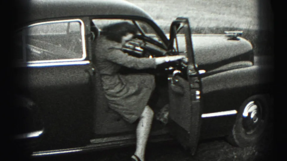 The woman steps into the driver's seat of the dark sedan, with the open door and dashboard visible in black-and-white 8mm footage.