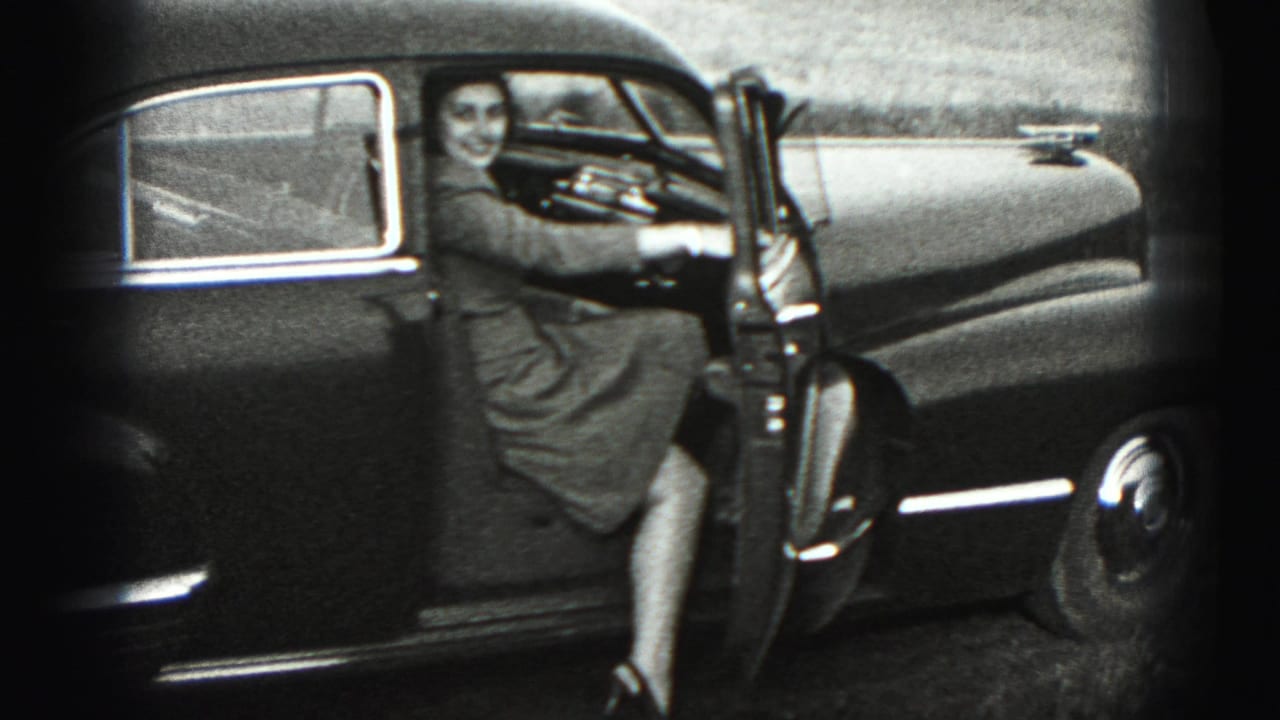 In Harrisburg-area 1975 home-movie footage, a woman stands beside an open older sedan door and smiles toward the camera.