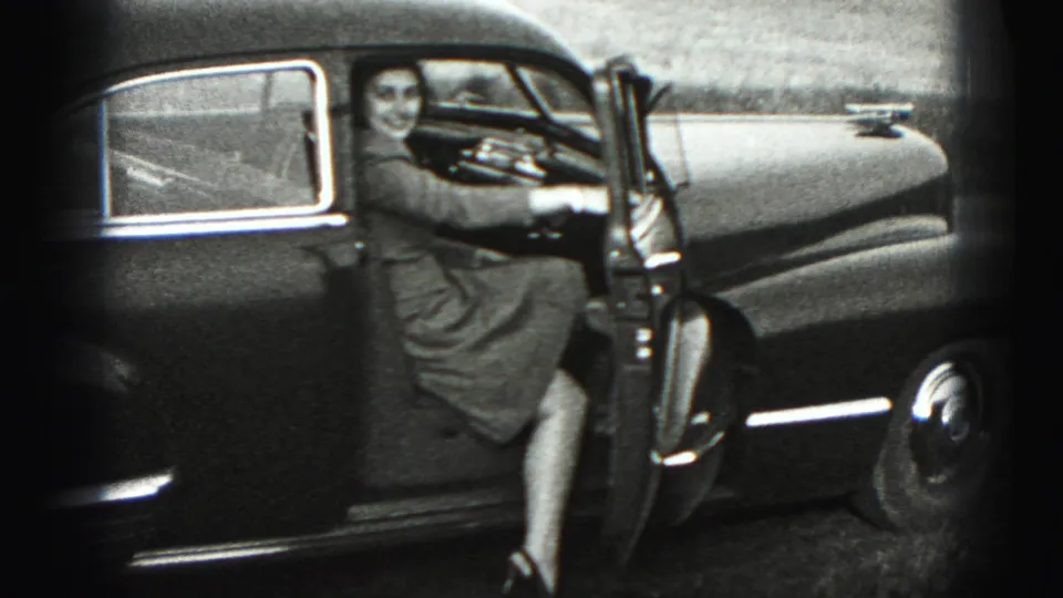 In Harrisburg-area 1975 home-movie footage, a woman stands beside an open older sedan door and smiles toward the camera.