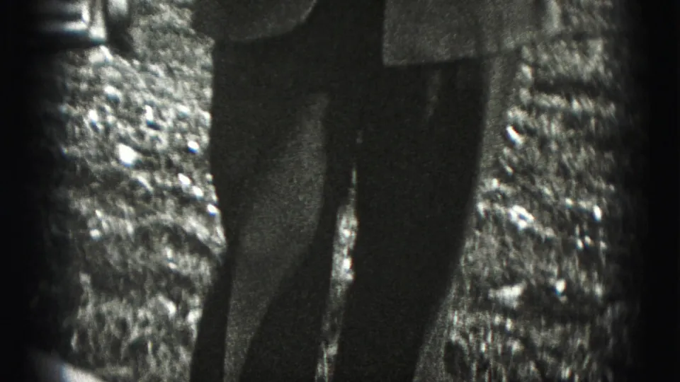 The handheld camera drops to the lower body of the uniformed subject, showing dark trousers and rough ground in the 1975 Harrisburg setting.