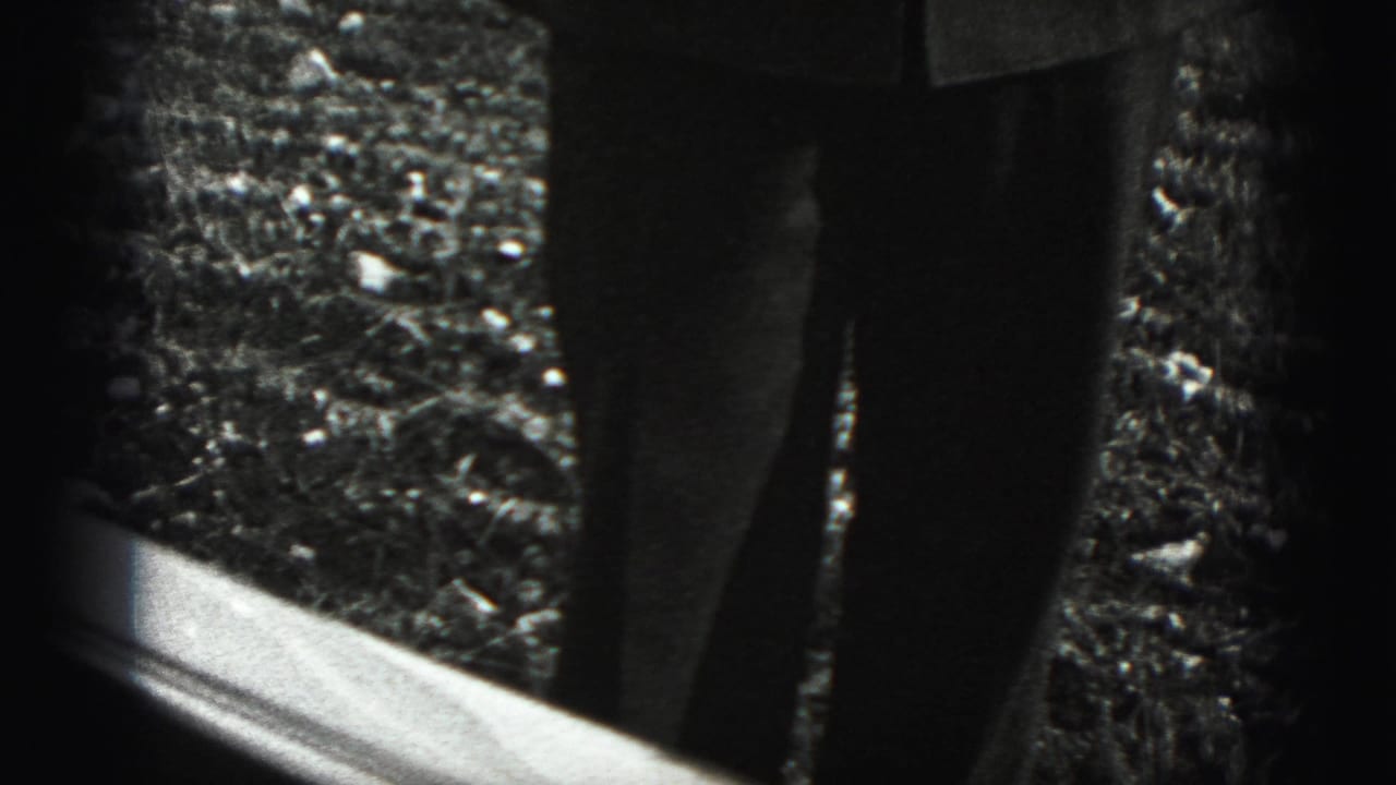 A cropped outdoor view shows trouser legs and gravel beside a reflective edge in the Harrisburg 1975 home-movie clip.
