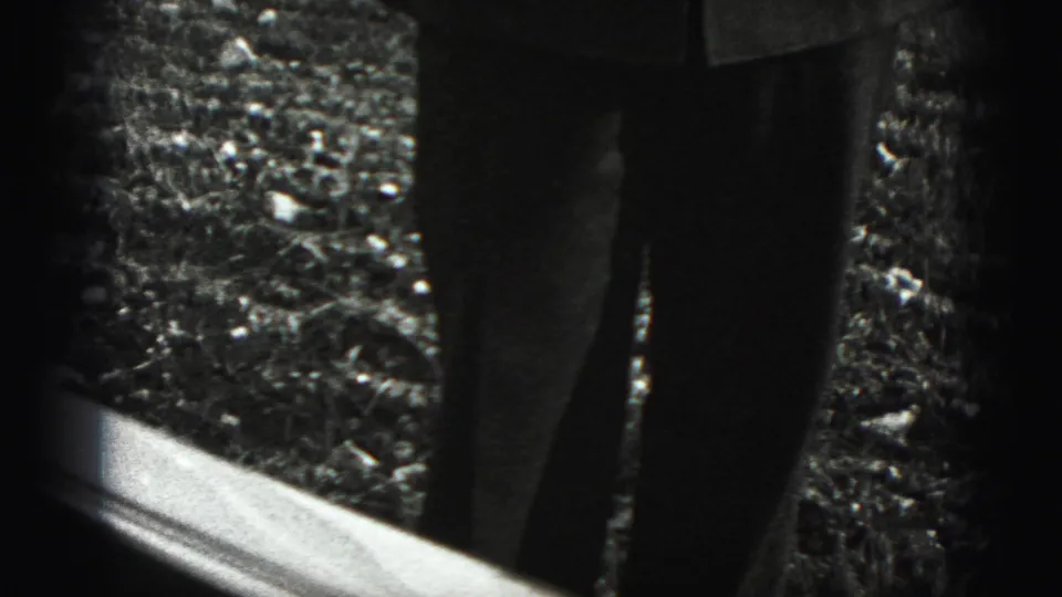 A cropped outdoor view shows trouser legs and gravel beside a reflective edge in the Harrisburg 1975 home-movie clip.