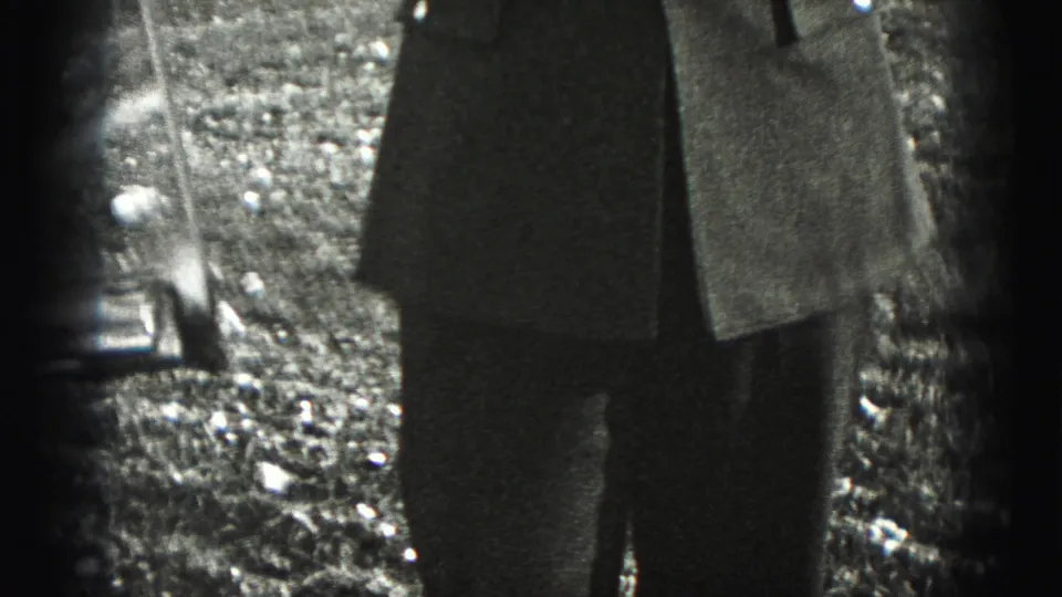 The lower half of a man in a dark military-style jacket appears outdoors on gravel in the 1975 Harrisburg footage.
