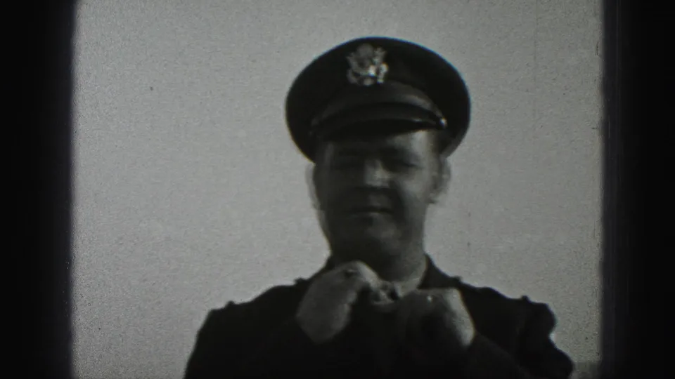 A uniformed adult man in a peaked cap appears to adjust his tie in the 1975 Harrisburg 8mm footage.
