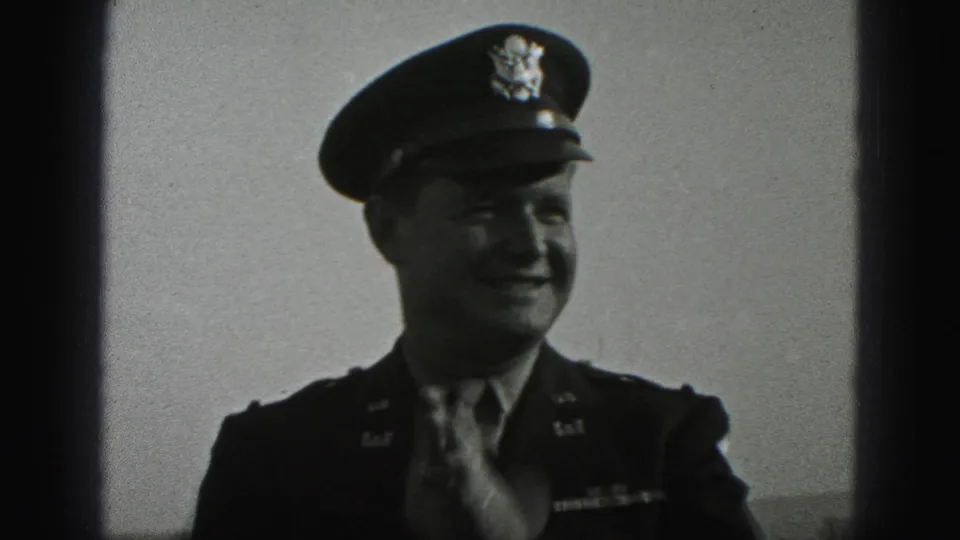 In 1975 Harrisburg home-movie footage, a uniformed soldier in a peaked cap smiles toward the camera while adjusting his tie.