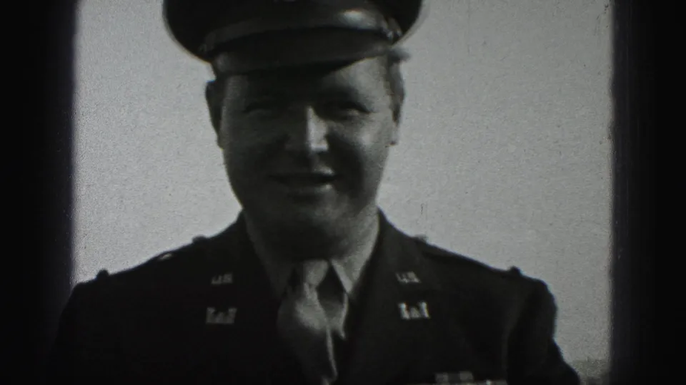 A soldier in dress uniform faces the camera in black-and-white 8mm footage, with cap, tie, and chest ribbons visible.