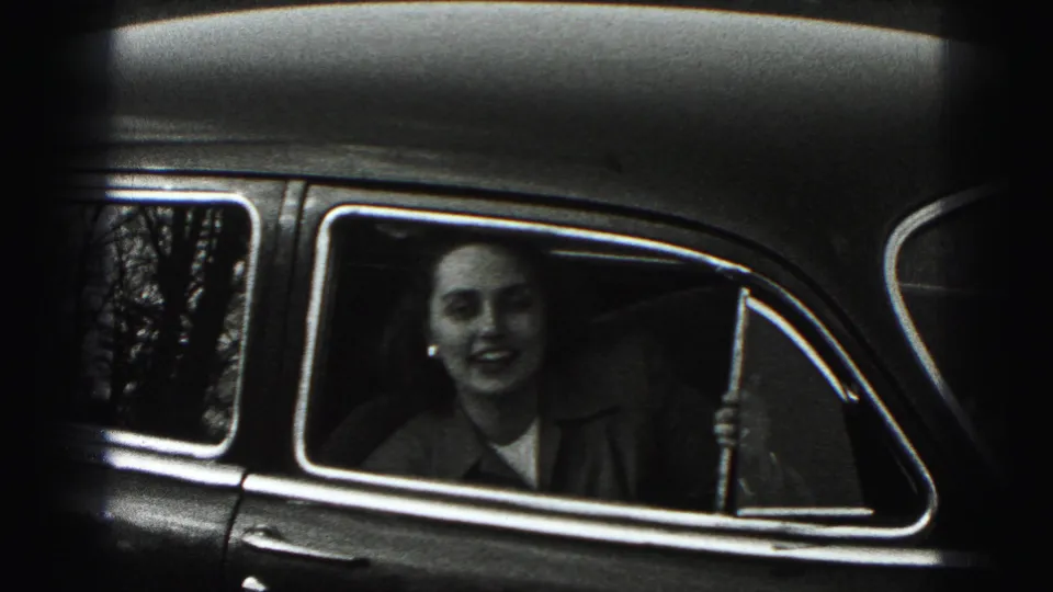 A woman seated inside a car smiles through the side window, with chrome trim and outdoor reflections visible.