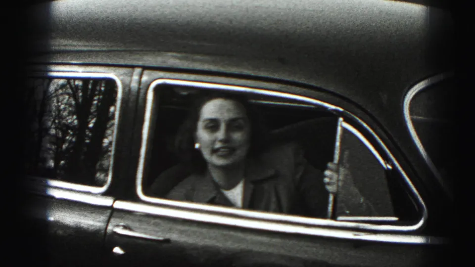 The woman remains seated inside the car, looking toward the camera through the window in grainy black-and-white 8mm footage.