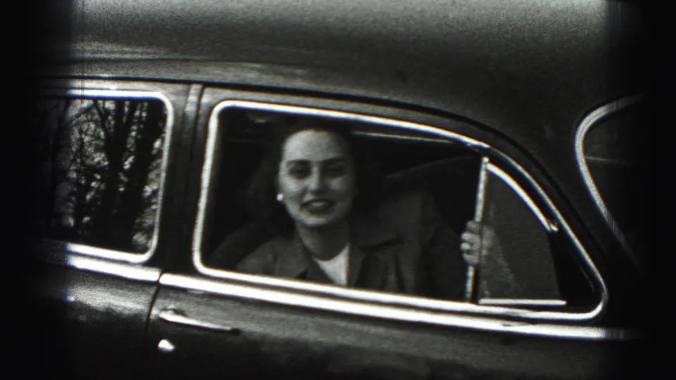 A woman in 1975 Harrisburg metadata context looks out from the passenger side of a dark vintage car, with trees reflected in the glass.