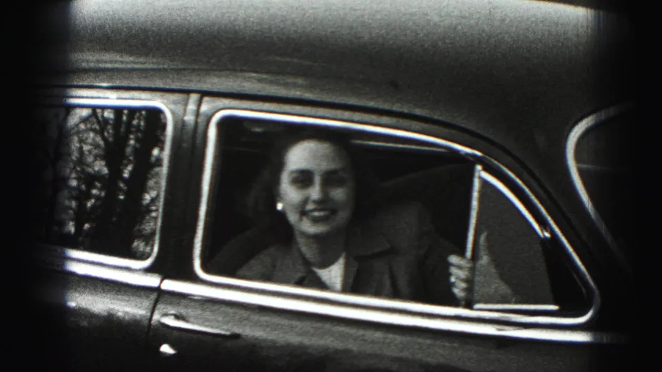 The woman appears to smile or speak from the passenger seat of a vintage car, with chrome trim and soft 8mm grain visible.
