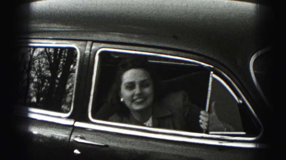 A woman in a collared jacket appears to laugh or speak through the open passenger window of a vintage car in 1975 metadata context.