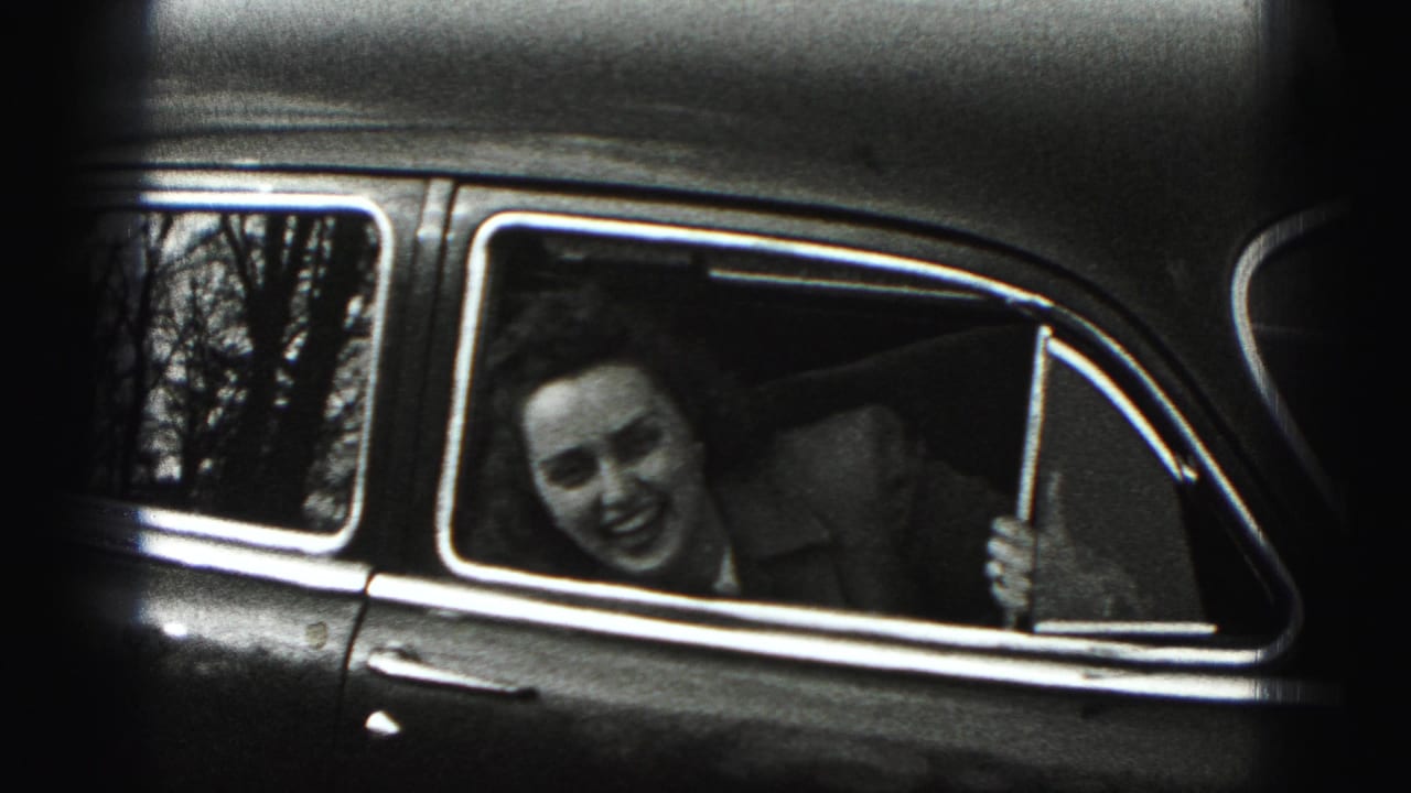 In 1975 Harrisburg home-movie footage, a smiling woman leans from the rear side window of an older car with trees reflected behind her.