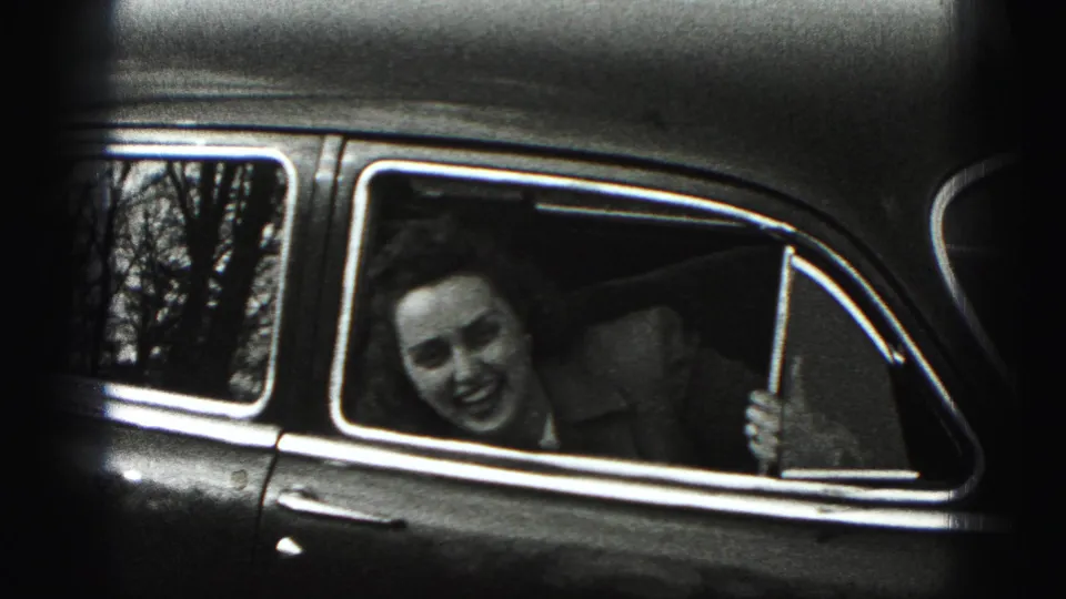 In 1975 Harrisburg home-movie footage, a smiling woman leans from the rear side window of an older car with trees reflected behind her.