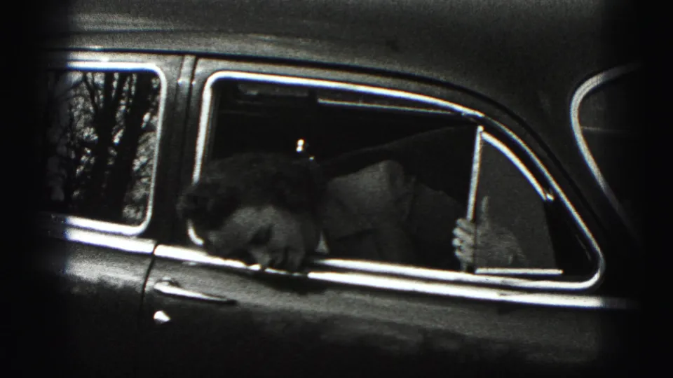 The woman ducks or bends inside the rear side window of the older car, with the seat and bright window trim visible.