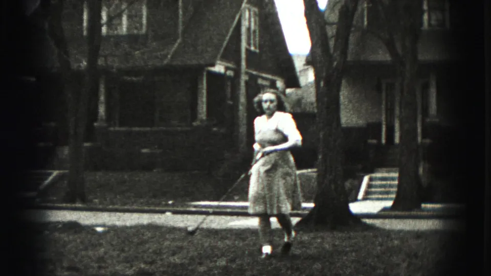 The woman walks across the Harrisburg yard holding a golf club, with houses, bare trees, grass, and sidewalk in the background.