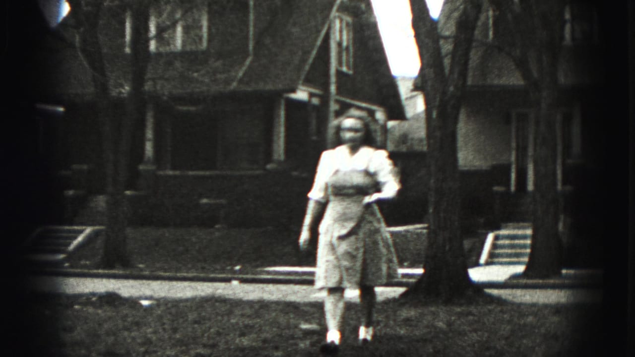 Woman in a light dress and apron walks across a grassy yard holding a golf club.