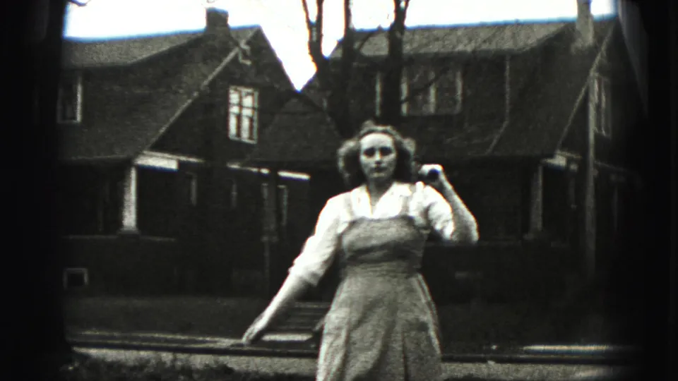 The woman approaches the camera with a golf club over one shoulder, framed by a grassy yard, bare trees, and residential houses.