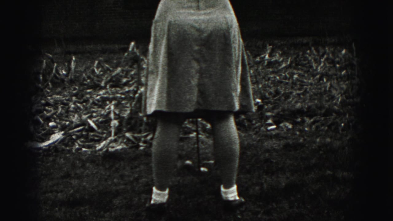 In 1975 Harrisburg home-movie footage, a woman stands from behind with a golf club near garden beds and a brick residence.