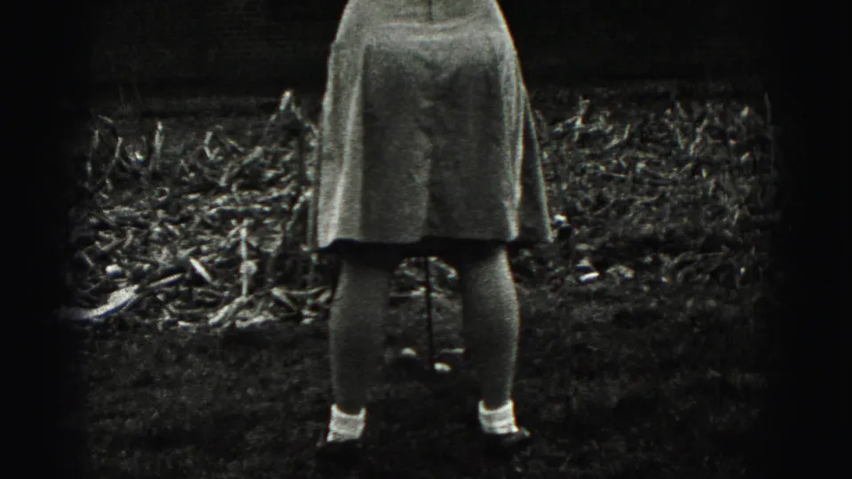 In 1975 Harrisburg home-movie footage, a woman stands from behind with a golf club near garden beds and a brick residence.