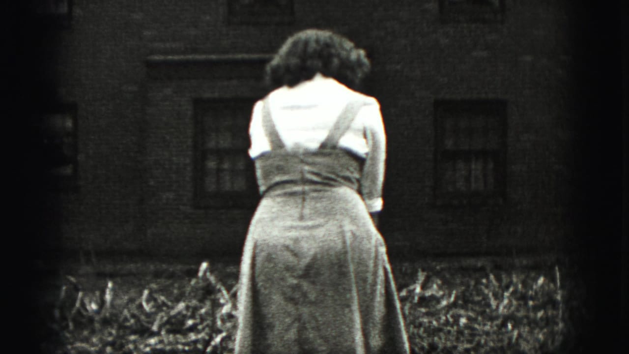 Woman stands in a backyard facing away near a brick building and garden bed.