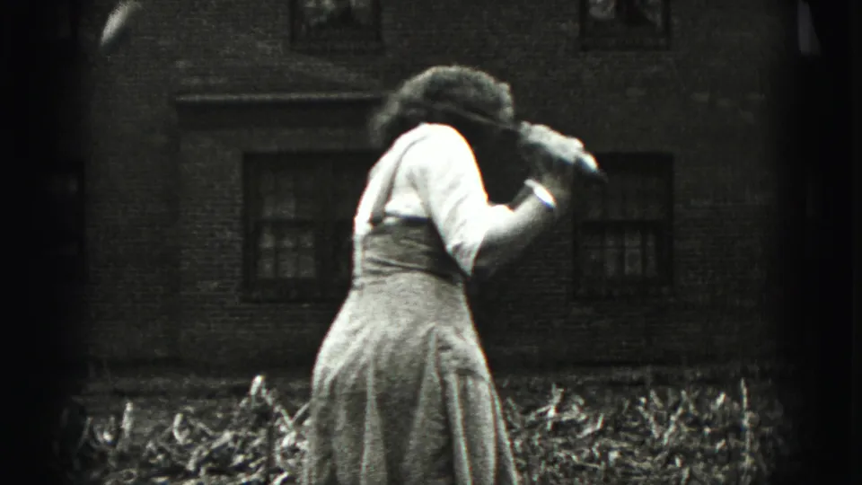 The woman bends forward in the yard, partly turned away from the camera, near low plants and a brick residential wall.