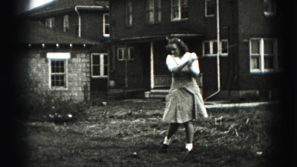 A woman in a dress finishes a backyard golf swing in 1975 Harrisburg home-movie footage.