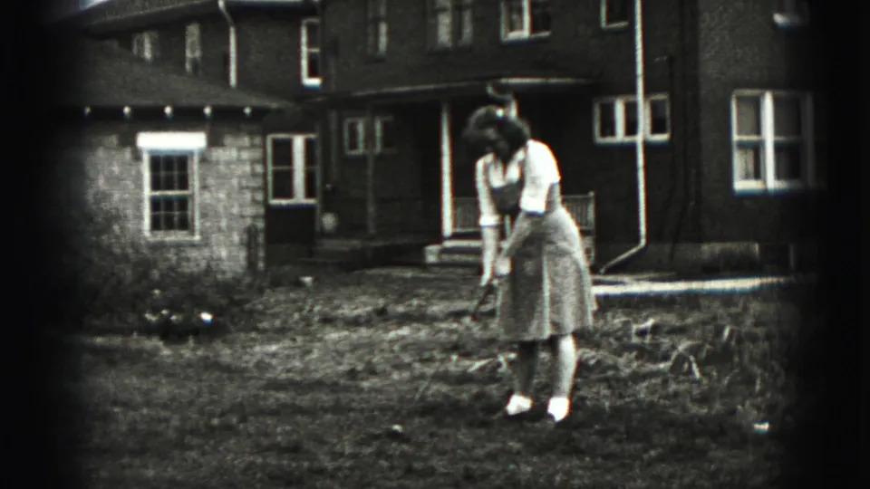 In Harrisburg 1975 home-movie footage, a woman prepares to hit a golf ball in a yard outside a multi-story house.