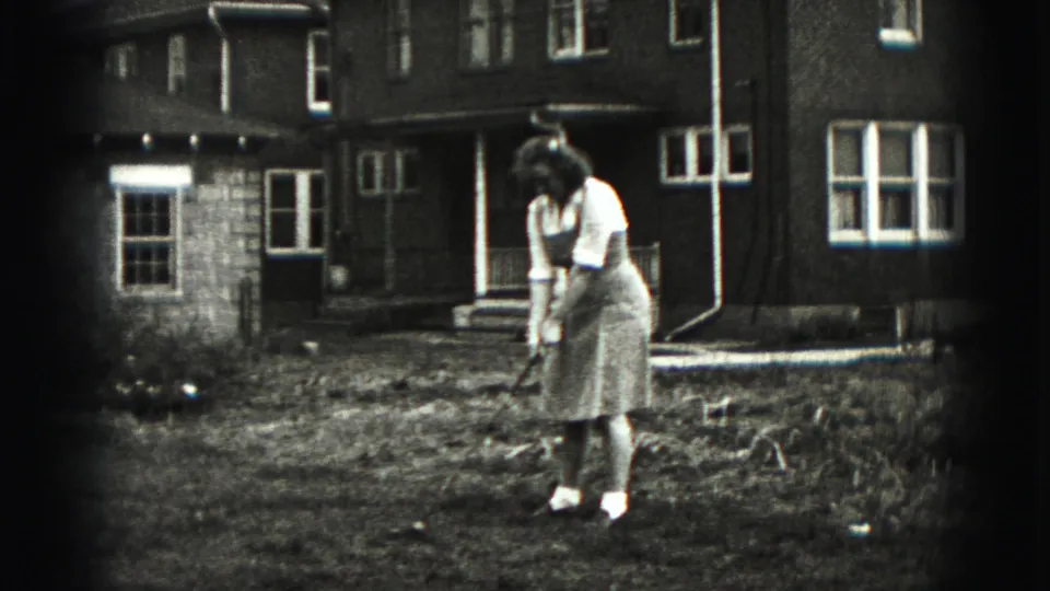 The woman remains focused on the golf ball or ground target while standing outside a Harrisburg house in 1975 footage.