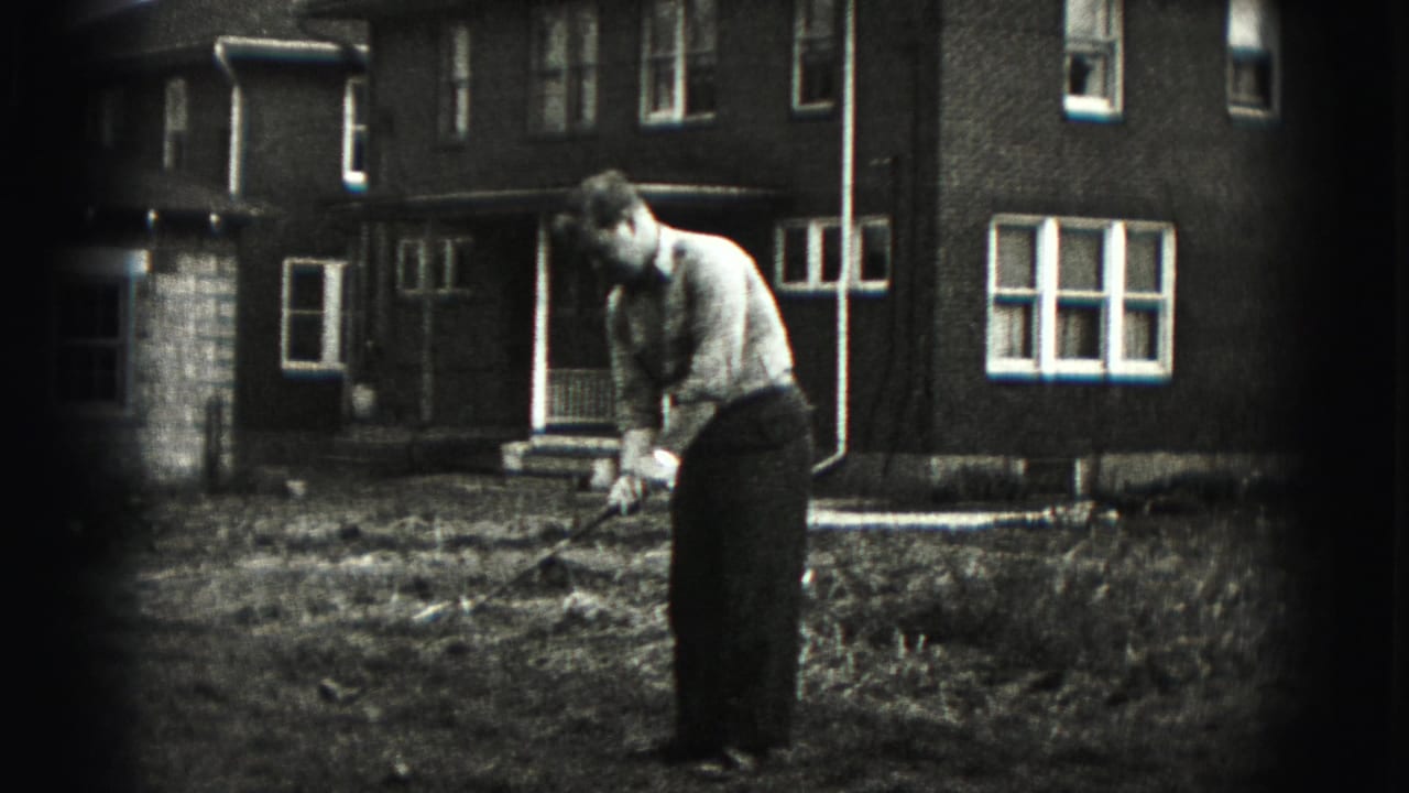 Man addressing a golf ball in a grassy yard outside a brick house.