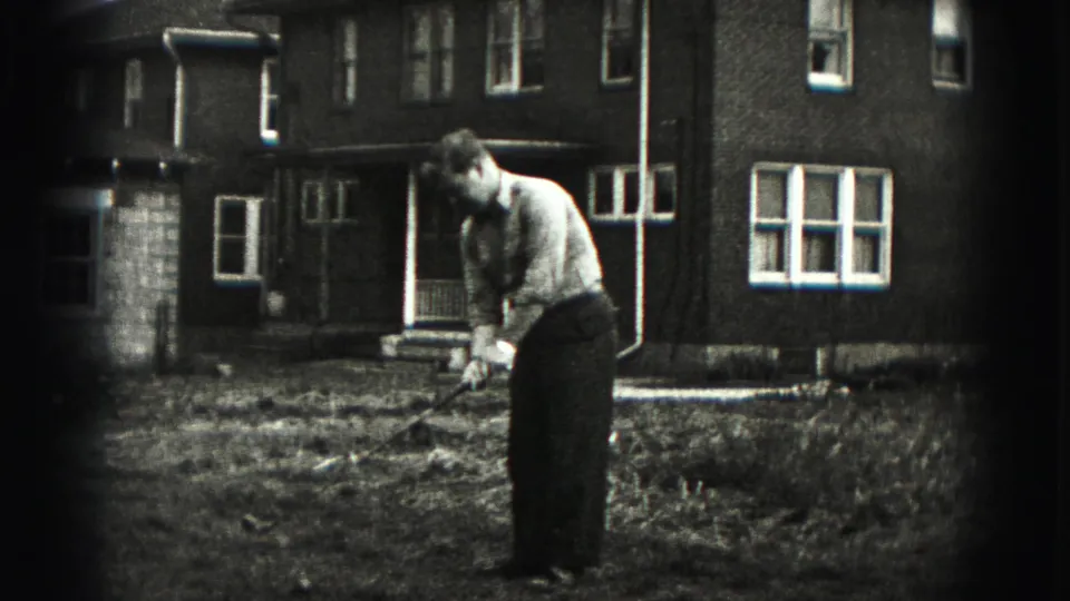 In 1975 Harrisburg home-movie footage, a man in a light shirt stands with a golf club in a grassy yard before a brick house.