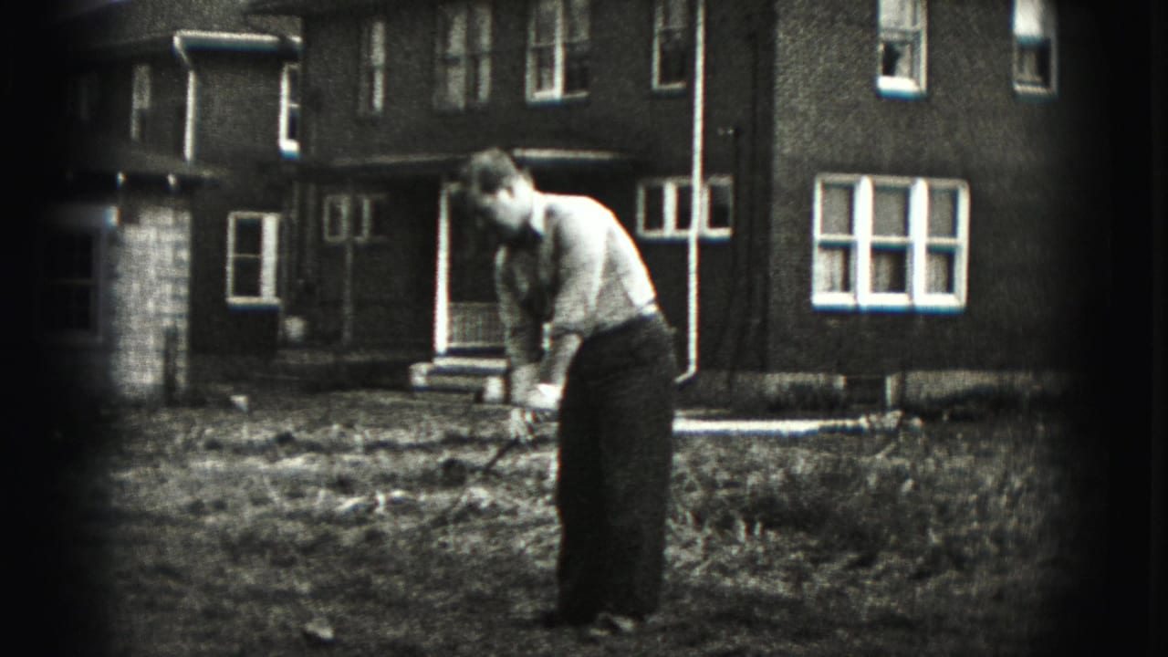 The golfer leans forward over the club in the Harrisburg yard, with a brick residential building and white-framed windows behind him.