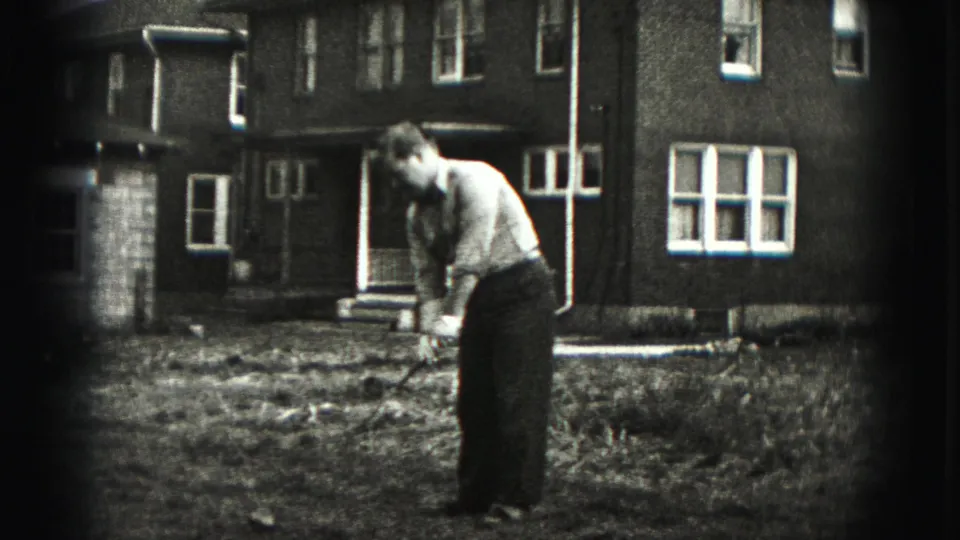 The golfer leans forward over the club in the Harrisburg yard, with a brick residential building and white-framed windows behind him.