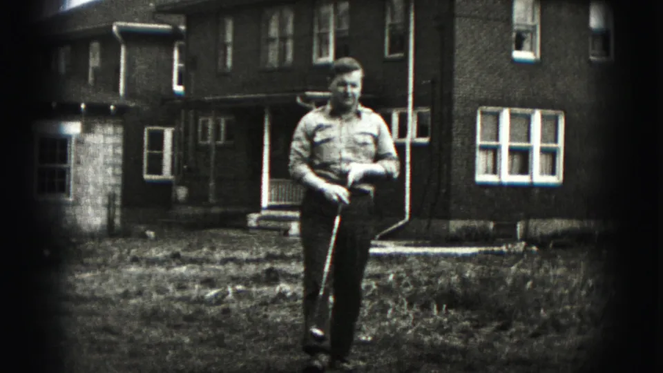 A man in a button-up shirt and dark trousers walks toward the camera with a golf club in a Harrisburg yard.