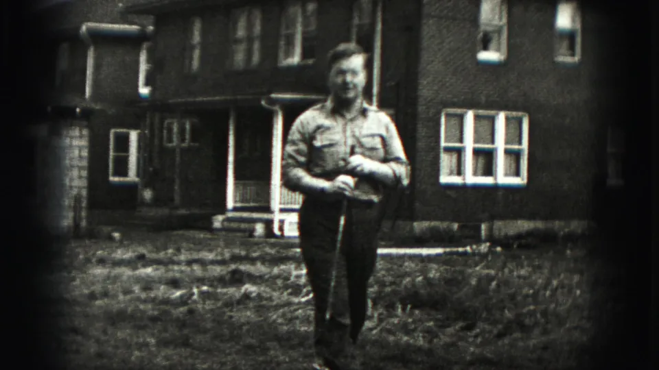 The man pauses near the camera with a golf club, framed by a grassy yard and brick residential exterior in 1975 footage.