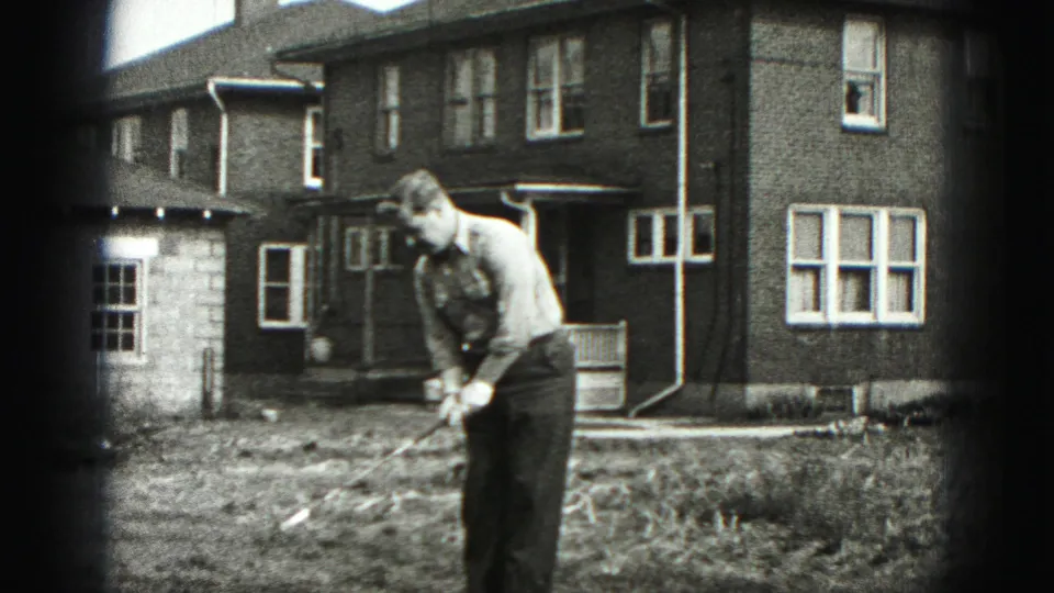 A man bends into a golf stance with the club lowered in a grassy yard beside a brick house in Harrisburg home-movie footage.