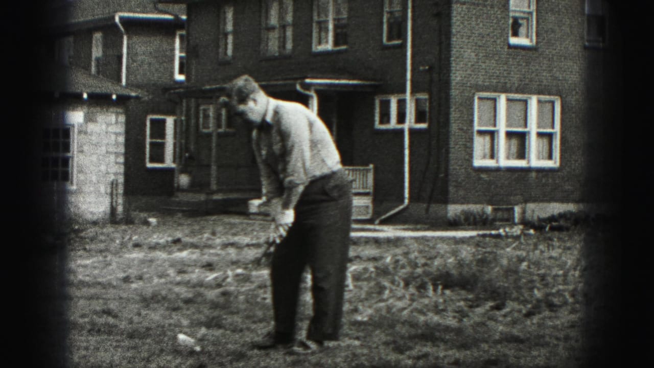 Man bends over a golf club in a grassy yard beside a brick house.