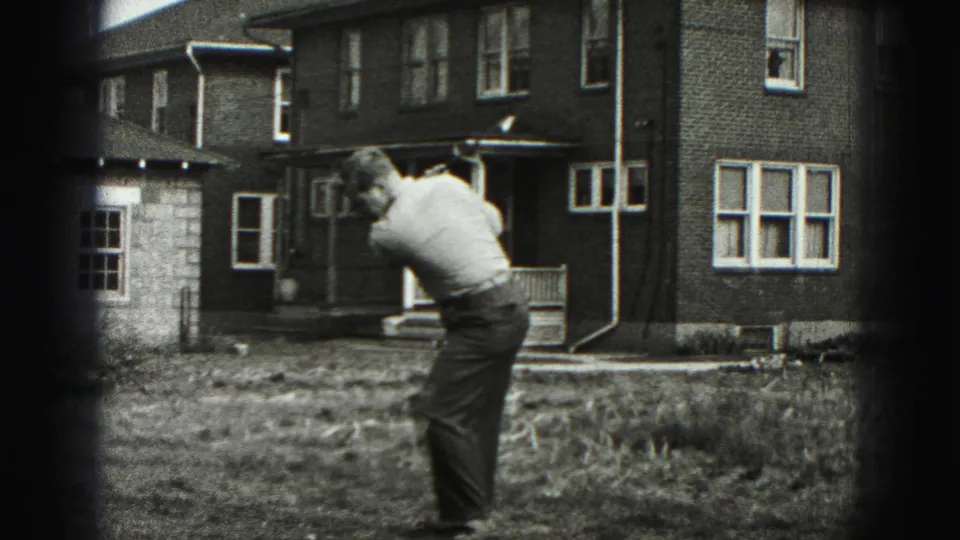 A man follows through on a golf swing in a grassy Harrisburg yard, with a brick house and porch behind him.