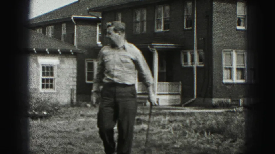 The man moves through the yard with a golf club beside a brick residential building in the 1975 Harrisburg footage.