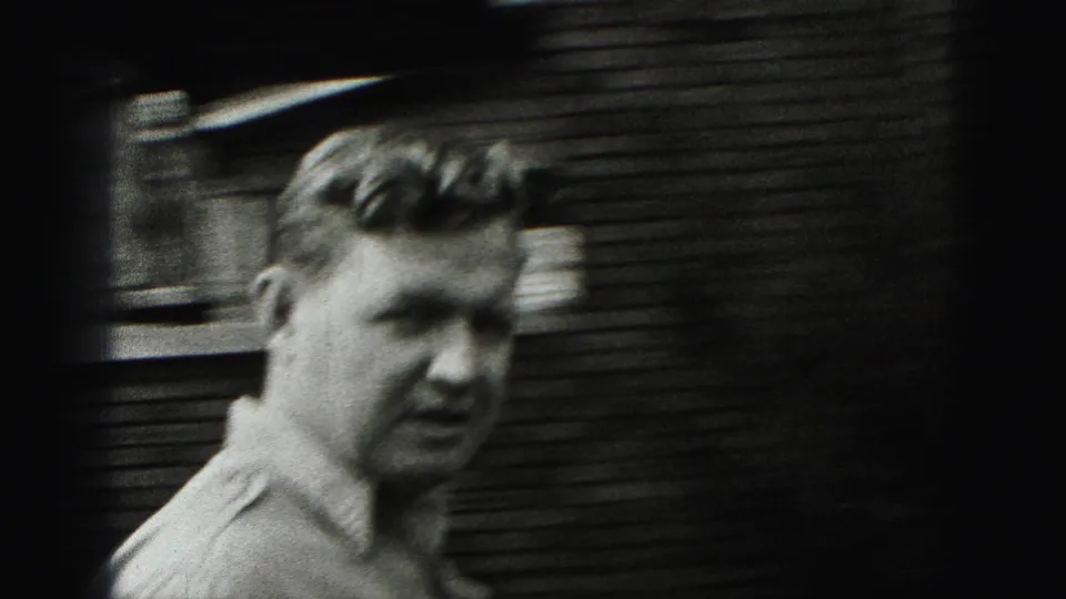 A close, soft-focused frame shows the man's face and light shirt near a dark exterior wall in the 1975 home movie.