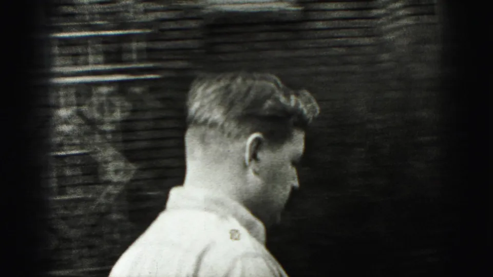 In 1975 Harrisburg home-movie footage, a young man in a light collared shirt passes a brick wall in profile.