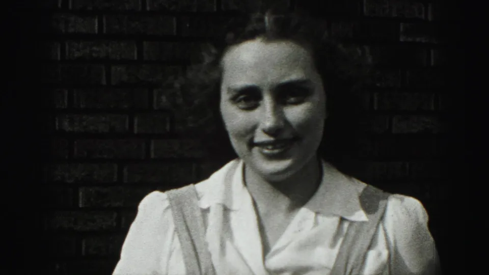The young woman smiles softly toward the camera, her white blouse and suspenders visible against the brick wall.