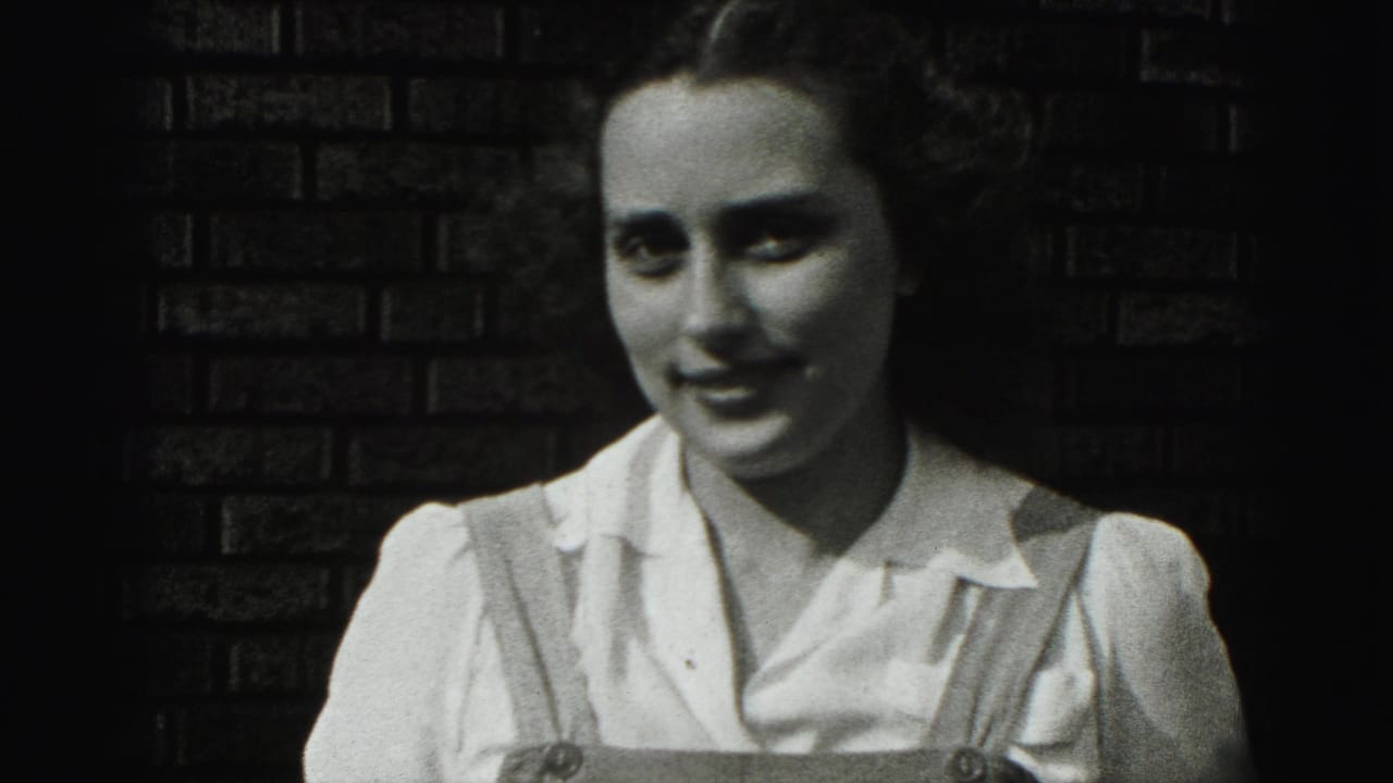 In 1975 Harrisburg home-movie footage, a young woman in a white blouse and overall straps smiles beside a dark brick wall.