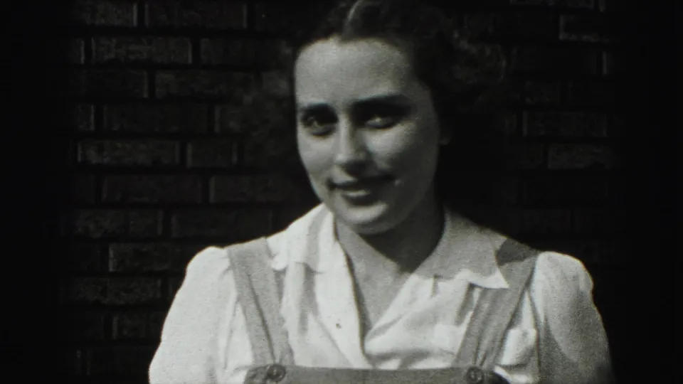 In 1975 Harrisburg home-movie footage, a young woman in a white blouse and overall straps smiles beside a dark brick wall.
